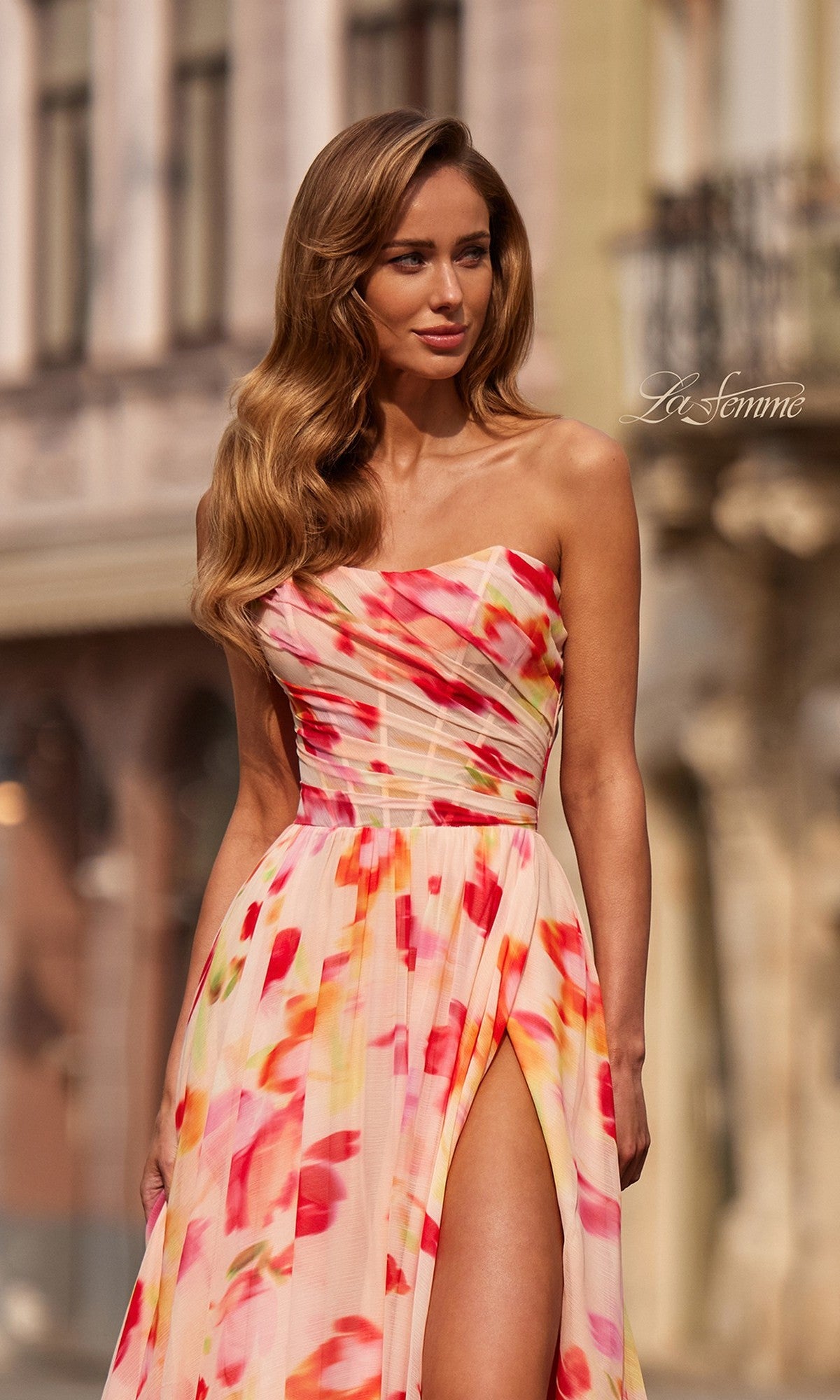 A woman stands outdoors in an urban setting wearing the La Femme 33404 long prom dressโa strapless, multicolored floral chiffon gown with a ruched corset bodice and high slitโher dress catching the light as she looks to the side.